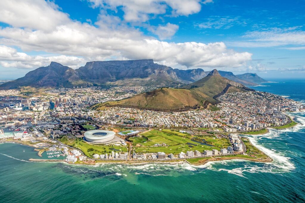 Number 4 of the top 5 travel destinations, an aerial view of Cape Town, South Africa, showcasing the city nestled between the iconic Table Mountain and the shimmering blue waters of the Atlantic Ocean.