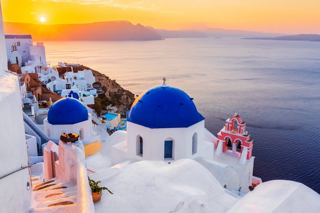 A breathtaking view of Santorini, Greece, at sunset, featuring whitewashed buildings with blue domes cascading down a cliffside overlooking the sparkling Aegean Sea.